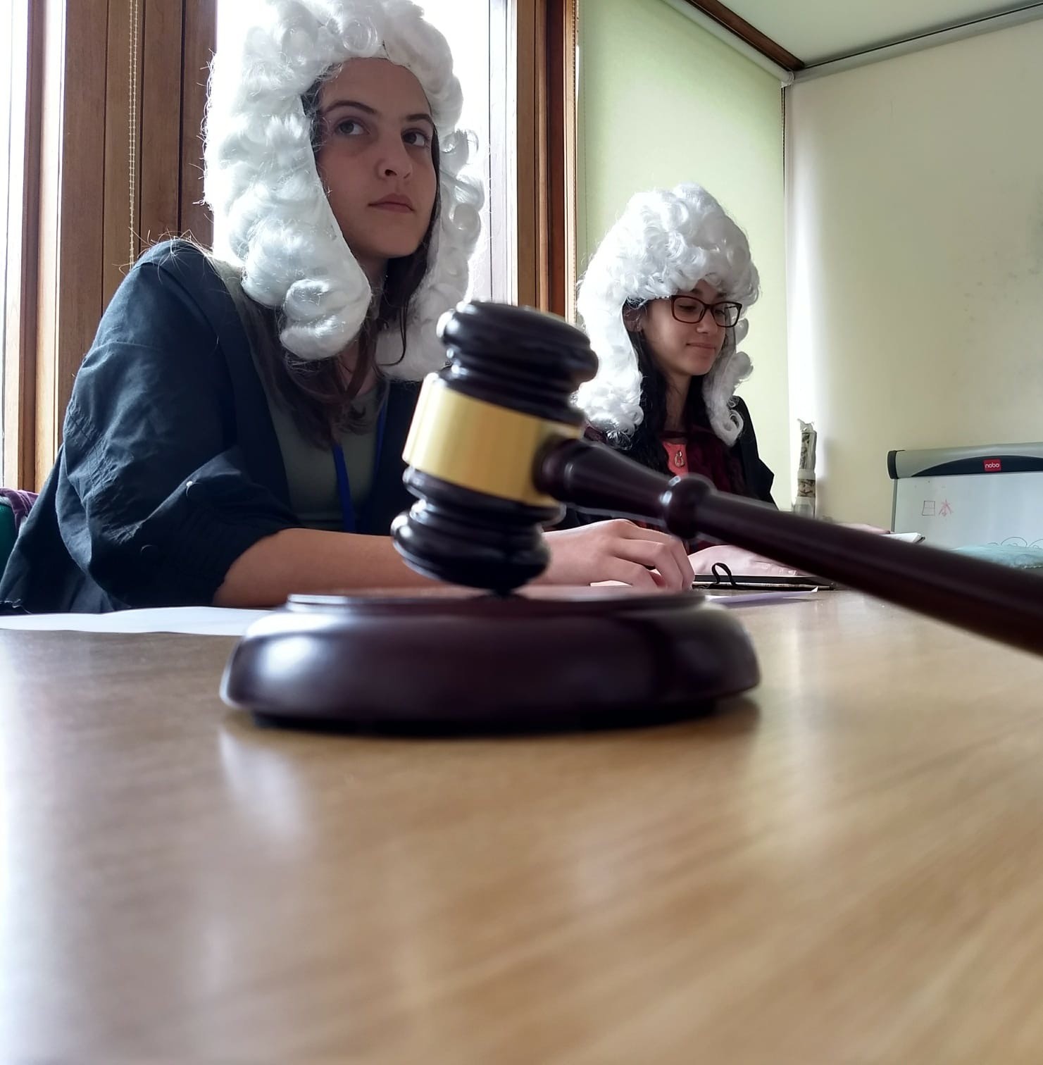 Two students sit at a classroom table, wearing fancy-dress judge wigs, in the background. In the foreground is a toy gavel resting on the table.