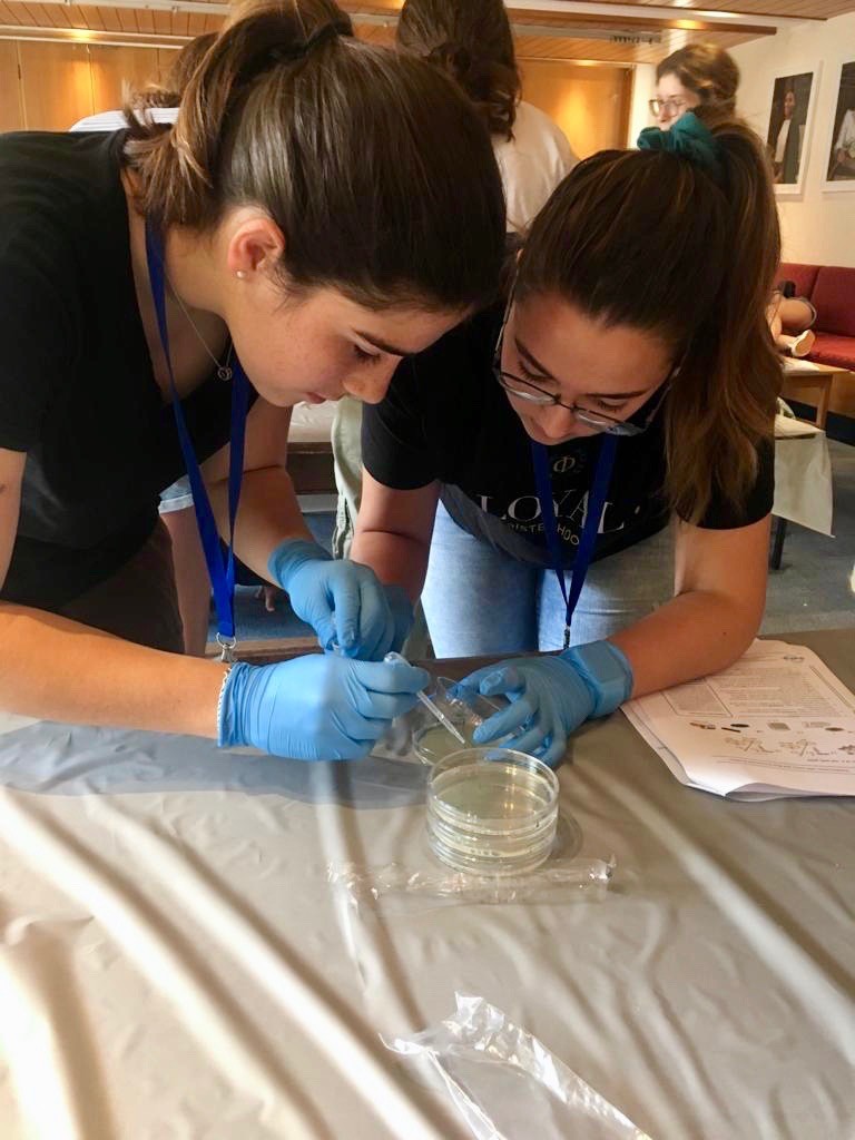 Two female students lean over a Petri dish. They are wearing blue rubber gloves and are bent over the table, getting a close look at their experiment.